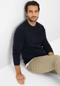 Granatowy sweter dziany z okrągłym dekoltem i ściągaczami na rękawach, w połączeniu z beżowymi spodniami. Siedząca pozycja przy białej ścianie.