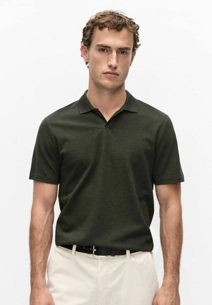 Jonge man met krullend haar die een donkergroen poloshirt met korte mouwen en lichtbeige broek met een zwarte riem draagt, staand tegen een witte achtergrond.