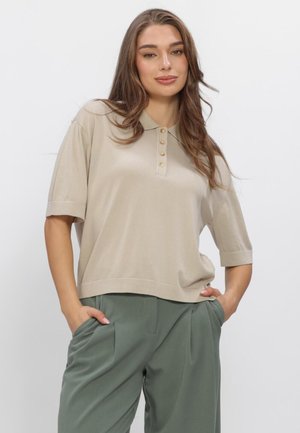 Młoda kobieta z długimi brązowymi włosami nosi beżową koszulkę polo z krótkim rękawem i luźne zielone spodnie, stoi z rękami w kieszeniach.