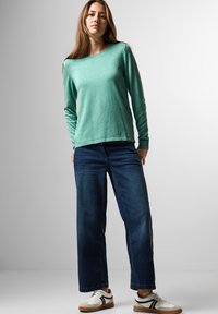 Mátově zelený top s dlouhým rukávem a kulatým výstřihem v kombinaci s tmavě modrými širokými džíny a bílými teniskami s černými pruhy.