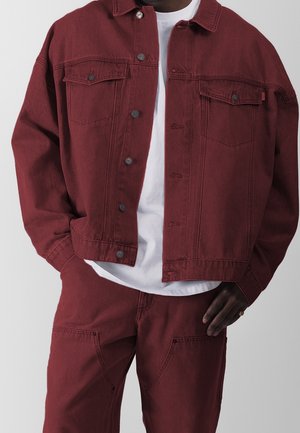 Homme portant une veste en jean bordeaux et un pantalon assorti sur un t-shirt blanc, une main dans la poche et l'autre reposant sur la hanche.