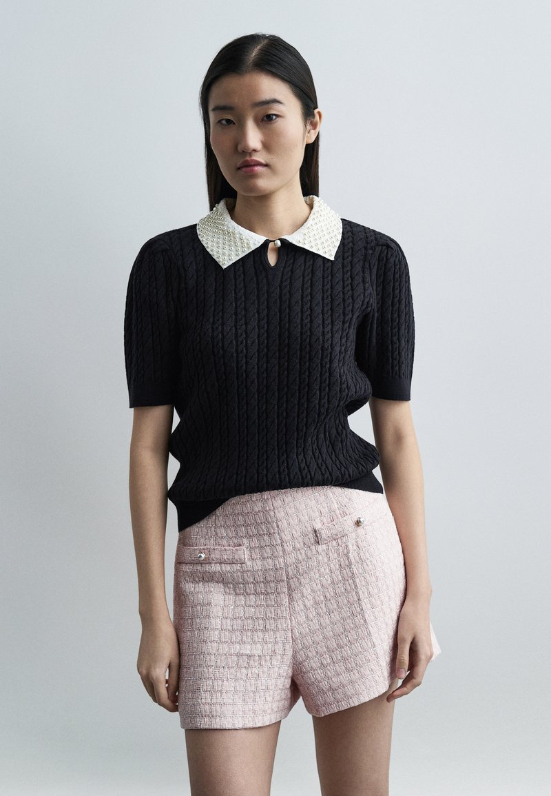 Jeune femme aux cheveux lisses et foncés portant un haut noir à manches courtes texturé avec un col blanc orné de perles et un short rose texturé.