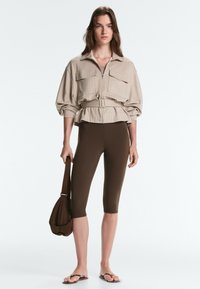 Femme portant une veste beige ceinturée et des leggings courts marron foncé, tenant un sac à main marron et portant des sandales tongs.