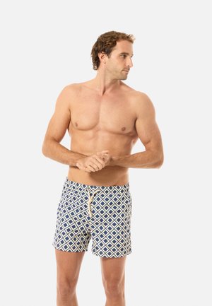 Oberkörperfreier Mann mit lockigem Haar, der blaue und weiße Schwimmshorts mit geometrischem Muster trägt, die Hände verschränkt und nach rechts blickt.
