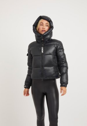 Mujer que lleva una chaqueta negra acolchada brillante con una capucha grande y pantalones negros ajustados, de pie sobre un fondo claro y liso.