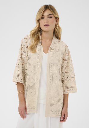 Lyshåret kvinde iført en beige åben-front hæklet cardigan med blomsterprint over en hvid top og vide bukser, accessoriseret med guld øreringe og armbånd.