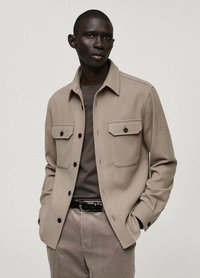 Giacca camicia in misto lana color beige con tasche frontali, chiusura a bottoni e vestibilità rilassata. Indossata sopra una maglietta a collo rotondo color taupe e pantaloni in velluto a coste.