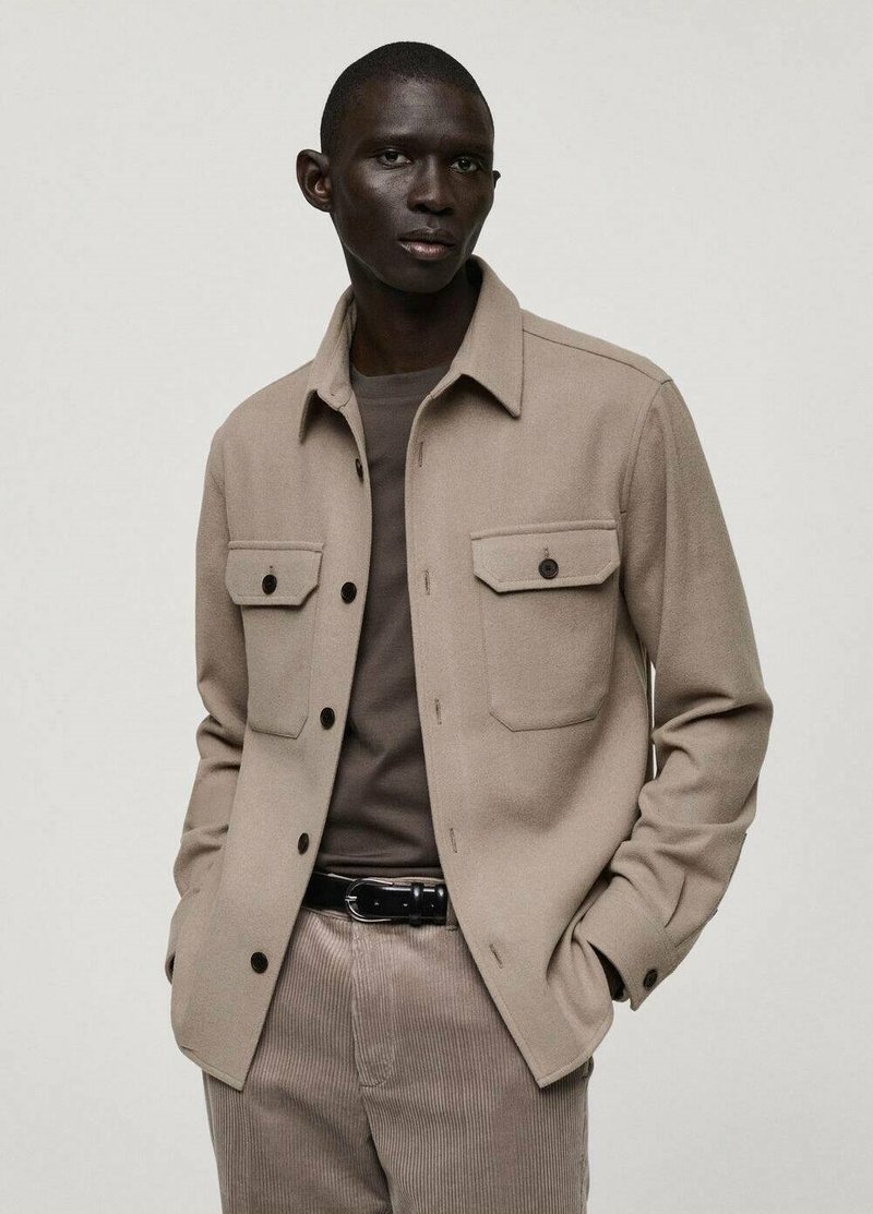 Giacca camicia in misto lana color beige con tasche frontali, chiusura a bottoni e vestibilità rilassata. Indossata sopra una maglietta a collo rotondo color taupe e pantaloni in velluto a coste.