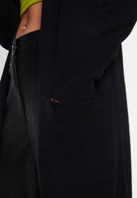 Czarny dziergany kardigan z prążkowanymi mankietami i przednią kieszenią, zestawiony z dopasowanymi czarnymi skórzanymi spodniami. Tekstura wydaje się miękka i przytulna.