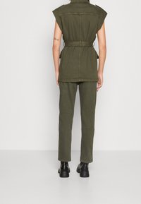 Combinaison vert olive à manches courtes avec taille ceinturée et poches latérales, assortie à un pantalon droit et des chaussures noires.