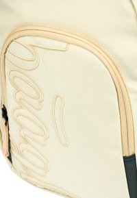 Zaino in tela beige texturizzata con zip crema, caratterizzato da un'evidente decorazione ricamata e un accentuazione nera nella parte inferiore.