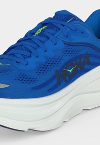 Μπλε αθλητικό παπούτσι HOKA με επάνω μέρος από δίχτυ, μαξιλαρωμένη λευκή σόλα, μπλε κορδόνια και πράσινες λεπτομέρειες λογότυπου στο πλάι και τη γλώσσα.