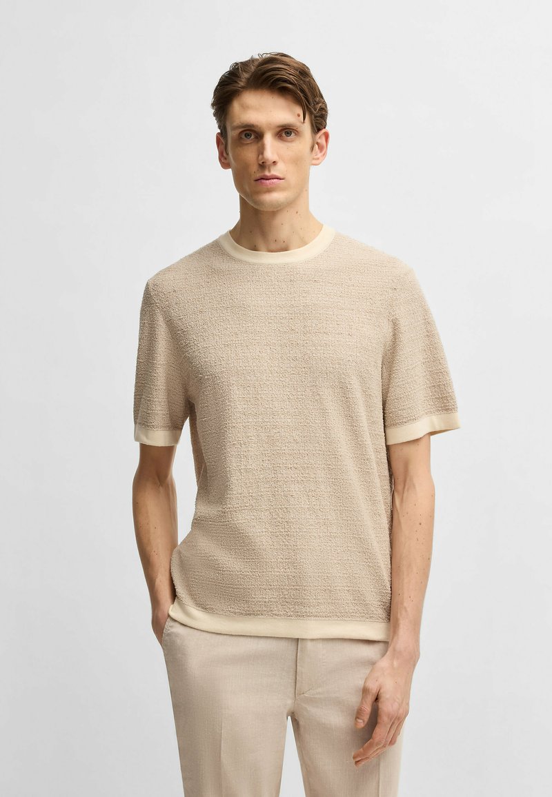 Camicetta beige a maniche corte con scollatura rotonda e rifiniture a contrasto di un beige chiaro. Il materiale sembra morbido con un motivo sottile.