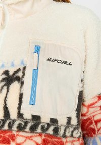 Pull en polaire crème avec des motifs rouges et bleus. Il possède une poche zippée avec des éléments en métal bleu et un logo "Ripcurl" brodé.