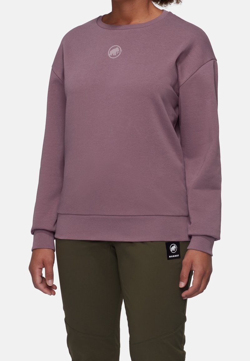 Femme portant un sweat-shirt mauve à manches longues et col rond ainsi qu’un pantalon vert olive avec des logos de la marque Mammooth sur la poitrine et la cuisse.