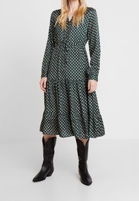 Groene gemusterde midi-jurk met lange mouwen, een knoopsluiting aan de voorkant en een gerimpelde zoom, gecombineerd met zwarte cowboylaarzen. Soepele stofstructuur.