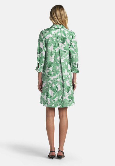 Frau steht, trägt ein knielanges weißes Kleid mit grünem Blumenmuster und dreiviertellangen Ärmeln, kombiniert mit schwarzen Riemchen-High Heels.