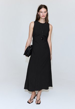 Femeie purtând o rochie midi neagră fără mâneci, tocuri negre cu barete, ținând o geantă plic neagră și purtând un colier lung negru cu pandantiv.