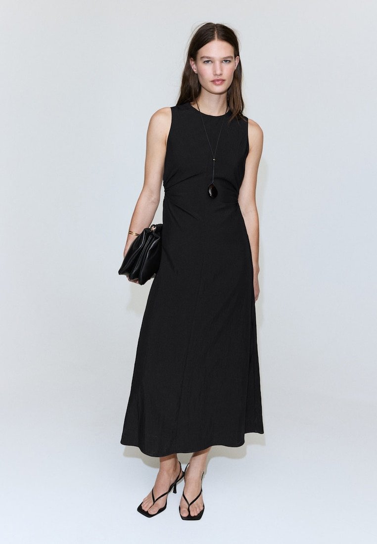 Femme portant une robe midi noire sans manches, des talons noirs à brides, tenant une pochette noire et portant un long collier pendentif noir.