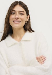 Witte, fluffy cardigan met een kraag, voorzien van knoopsluiting en geribbelde textuur. Lange mouwen met zachte randen, wat zorgt voor een gezellige uitstraling.
