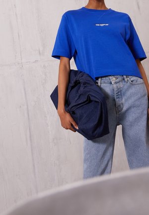 Osoba ubrana w niebieski krótki T-shirt i jasnoniebieskie dżinsy, trzymając dużą złożoną ciemnoniebieską torbę materiałową przy teksturowanej szarej ścianie.