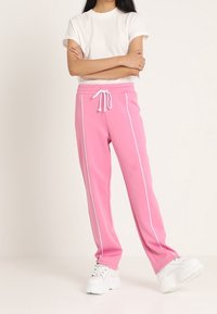 Pantalones de chándal rosas con rayas blancas en los lados y cinturilla con cordón, combinados con una camiseta blanca y zapatillas gruesas blancas. Textura suave.