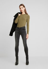 Blusa de manga longa verde-oliva combinada com calças skinny cinza-escuras e botas de cano curto pretas de salto, com uma peça de roupa exterior preta pousada sobre o ombro.