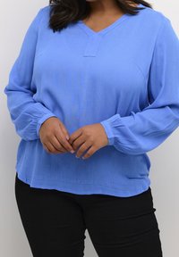 Blusa de manga larga azul claro con escote en V y tejido texturizado. Presenta puños fruncidos y un corte holgado. Combinada con pantalones negros.