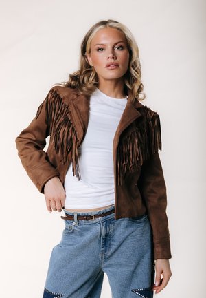 Kobieta z blond włosami ubrana w brązową zamszową kurtkę z frędzlami, białą koszulkę i niebieskie jeansy, pozująca na jednolitym jasnym tle.