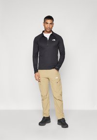 Zwarte lange mouwen, rits-top met logo, gecombineerd met lichtbeige cargobroek met zakken. Zwarte wandelschoenen maken de outfit compleet.