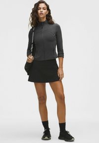 Mujer de pie con chaqueta negra con cremallera, falda negra, calcetines negros y zapatillas, sosteniendo una bolsa negra sobre su hombro.