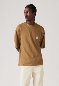 Tricou din bumbac maro, cu mâneci scurte, cu un buzunar ce are o etichetă, asortat cu pantaloni de culoare deschisă. Textură netedă, croi lejer.