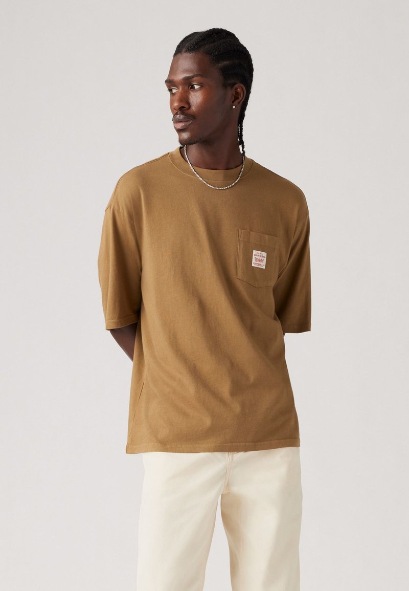 T-shirt in cotone marrone con maniche corte, tasca con etichetta, abbinato a pantaloni di colore chiaro. Tessuto morbido, vestibilità rilassata.