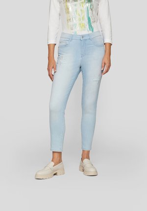 Hellblaues Denim-Jeans mit schlanker Passform, ausgestattet mit destroyed Details an der Vorderseite und im Knöchelbereich. Getragen mit einem weißen, bedruckten Oberteil und beigen Schuhen.