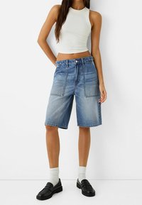 Hoge taille blauwe denim shorts met een lichte wassing. Heeft twee voorzakken en een knoopsluiting. Gecombineerd met een witte mouwloze top en zwarte instappers.
