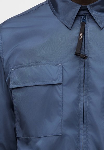 Gros plan sur une chemise bleue avec un col, une poche poitrine à rabat et une tirette de fermeture éclair noire portant la marque "ASPESI" sur le devant.
