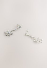 Srebrne naušnice s motivom pahuljica ukrašene brojnim prozirnim kristalima, prikazuju mješavinu glatkih i teksturiranih površina.