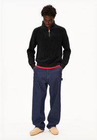 Mann trägt einen schwarzen Reißverschluss-Pullover, darunter ein rotes Shirt, weite blaue Jeans und beiger Mokassinschuhe, steht mit den Händen in den Taschen.