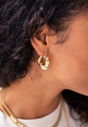 Kręcone ciemne włosy z nakręconą złotą obręczą na uchu oraz warstwowymi złotymi łańcuszkami na szyi na białym topie.