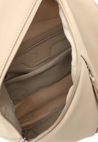 Beige ryggsäckens insida har en slät yta, flera fack och ett zippered-fack. Diskret logotyp präglad på foder.