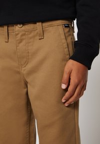 Khaki broek met een gladde textuur, riemlussen en een knoopsluiting. Hand rust op de dij, contrasterende zwarte trui zichtbaar erboven.