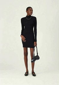 Sieviete, tērpusies melnā garo piedurkņu ribu turtleneck kleitā, tur melnu somu un valkā melnus kurpes ar sprādzi.