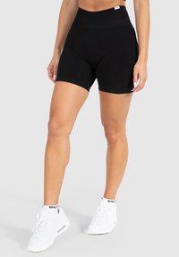Schwarze, hochtaillierte Fitnessshorts aus dehnbarem Material mit einer glatten Textur und Saum, kombiniert mit weißen Sportschuhen.
