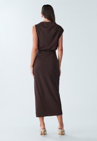 Robe marron sans manches avec une taille ajustée, longueur genou, tissu texturé, présentant un dos drapé, associée à des talons de couleur claire.