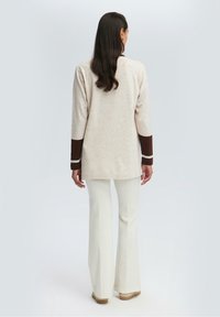 Top beige a maglia con maniche tre-quarti, polsini marrone scuro e pantaloni svasati di colore chiaro. Texture liscia e vestibilità comoda.