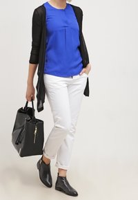 Sininen pusero laskostetuilla yksityiskohdilla, musta avoin neuletakki, valkoiset housut ja mustat nilkkurit, jotka pitävät teksturoitua mustaa laukkua.