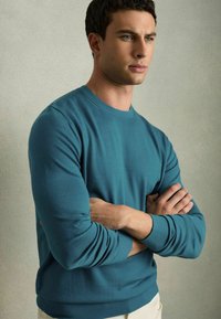 Zöld crew neck pulóver hosszú ujjakkal és bordás mandzsettákkal, amelyet egy model visel, karjait keresztbe tett állapotban, semleges háttér előtt.