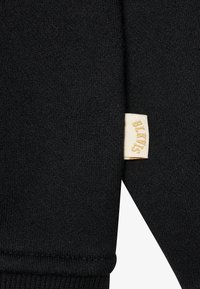 Tissu noir côtelé avec une surface texturée et une petite étiquette crème ornée de texte doré. Le design comprend un détail de bord côtes replié.