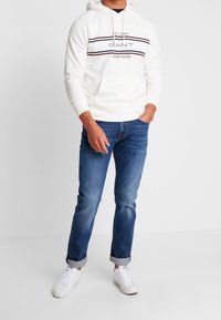 Felpa bianca con cappuccio e righe rosse e blu; logo "GANT NEW HAVEN" sul petto. Abbinata a jeans blu in denim e sneaker bianche.