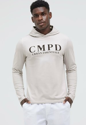 Young man wearing a light beige hoodie with "CMPD Urban Essentials" text, hands in pockets, standing against a plain light background.
